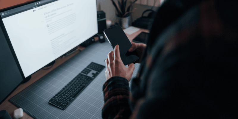 man looking at mobile phone while working on desktop