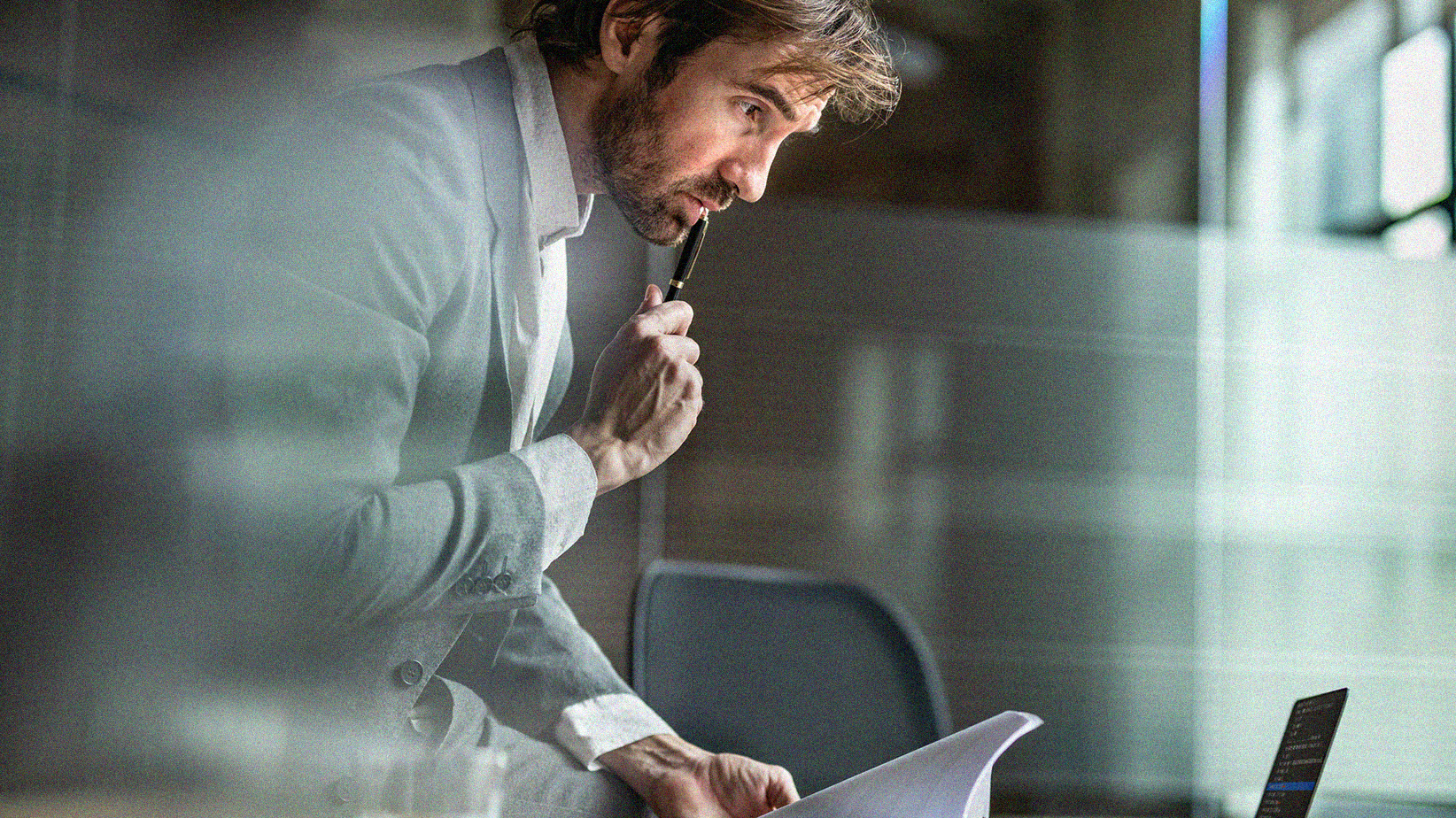 Business professional contemplating and analyzing paperwork and leaning over desk with a pen raised to his mouth