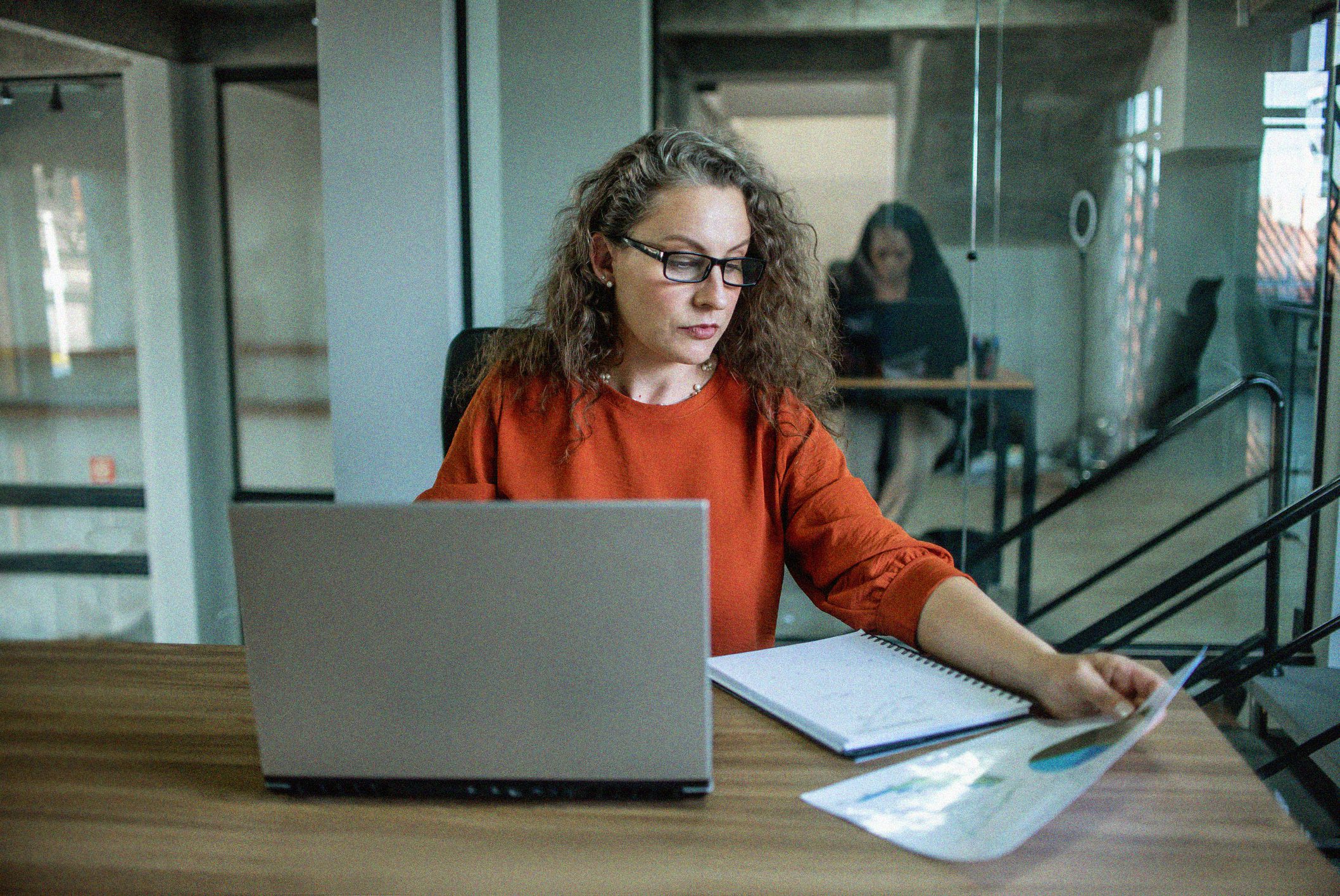 Finance professional, woman with glasses, at desk assessing a chart behind a laptop