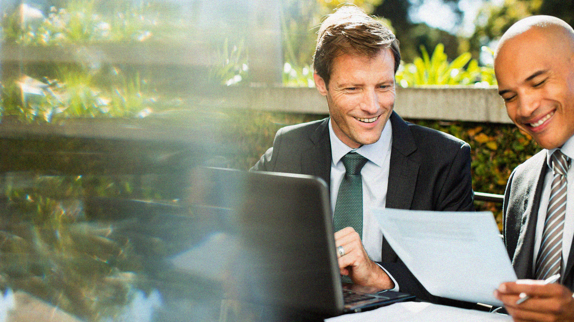 Two men in suits smiling and collaborating with paperwork and a laptop outdoors