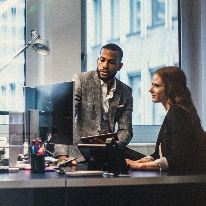 Two business professionals collaborate in an office setting at a desk discussing something viewed on a desktop computer.