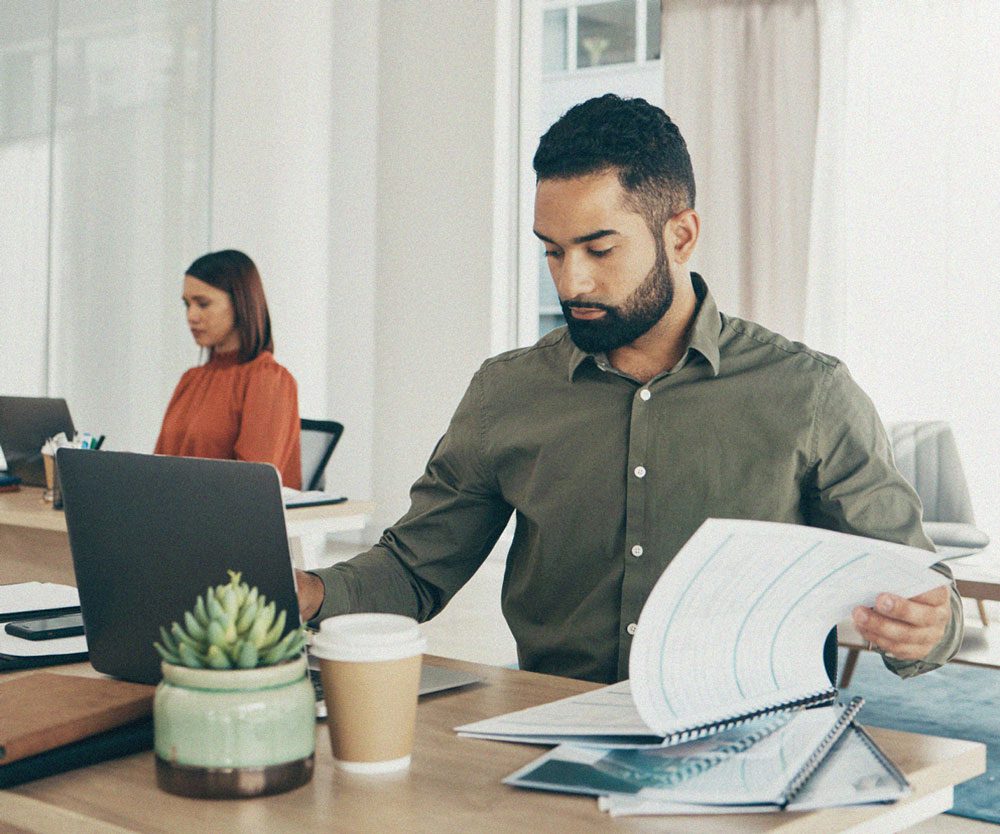 A bearded man in a green shirt is working at a modern office desk, focused on a spiral-bound report while using a laptop. A woman in a red blouse is seated at another desk in the background. The workspace is minimalistic, with neutral tones, a potted succulent, and a coffee cup on the desk.