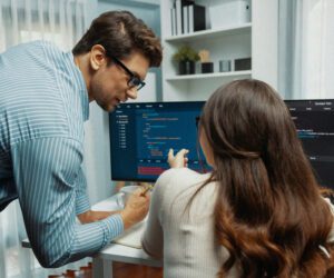 A man in a striped shirt leans over to assist a woman with long brown hair who is seated at a desk, pointing at a computer screen displaying code. They appear to be collaborating on a programming task in a modern, well-lit office with dual monitors and minimalistic decor.