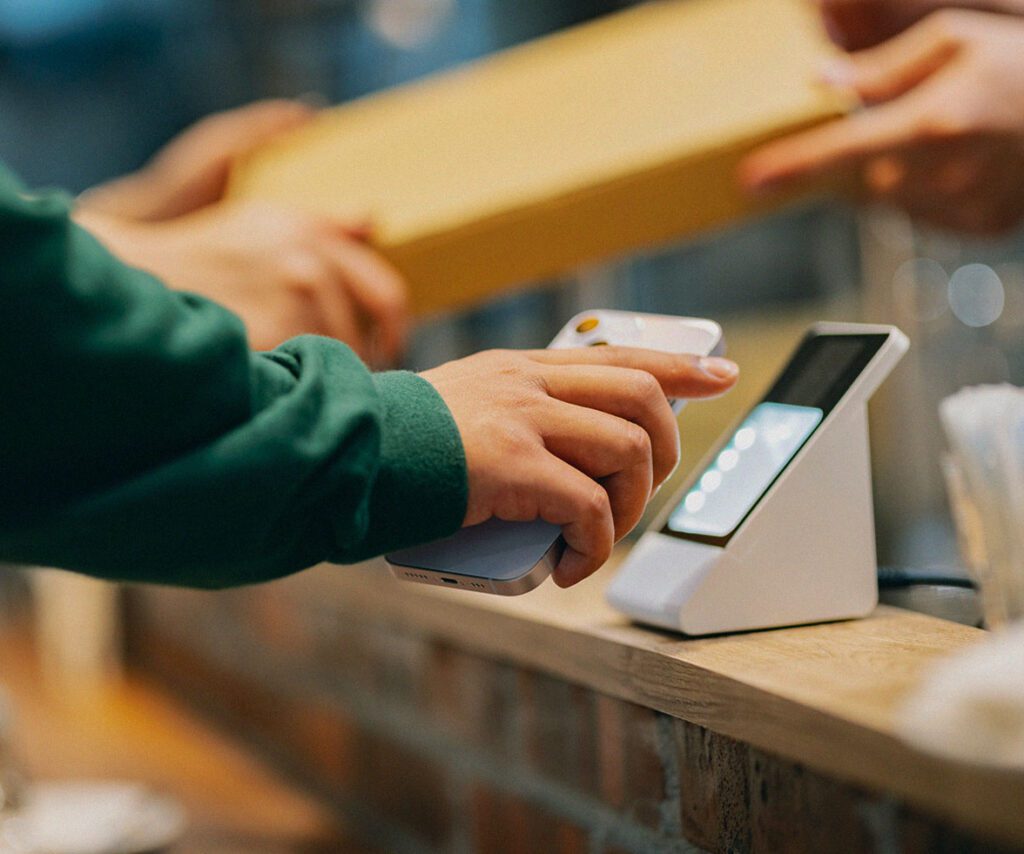 A person in a green sweatshirt makes a contactless payment using a smartphone at a modern point-of-sale terminal while another individual hands over a yellow package across the counter.