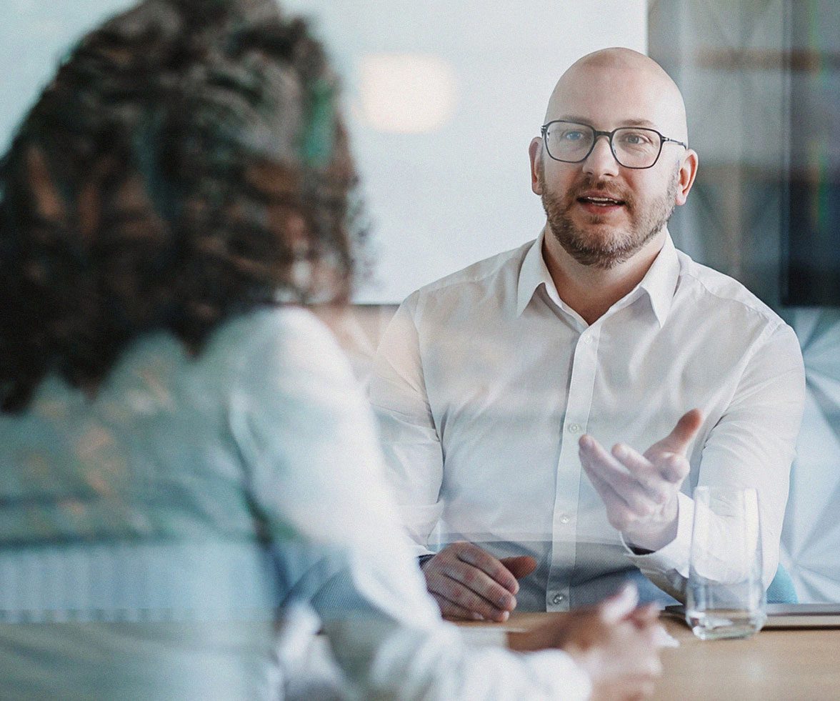 A bald man wearing glasses and a white shirt speaks while gesturing during a business meeting across the table from a woman with curly hair, seen from behind through a glass partition.