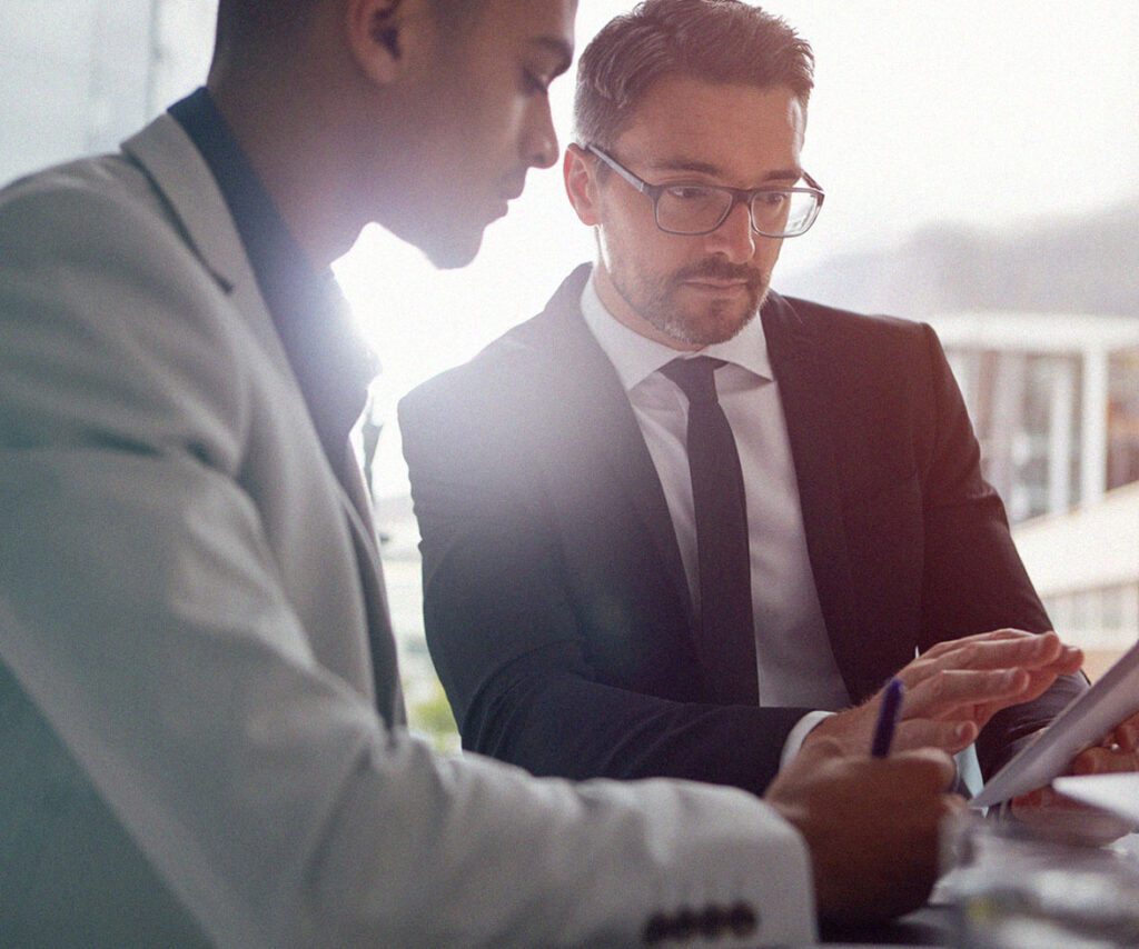 Two businessmen in suits having a focused discussion while reviewing information on a digital tablet in a bright, modern office setting.