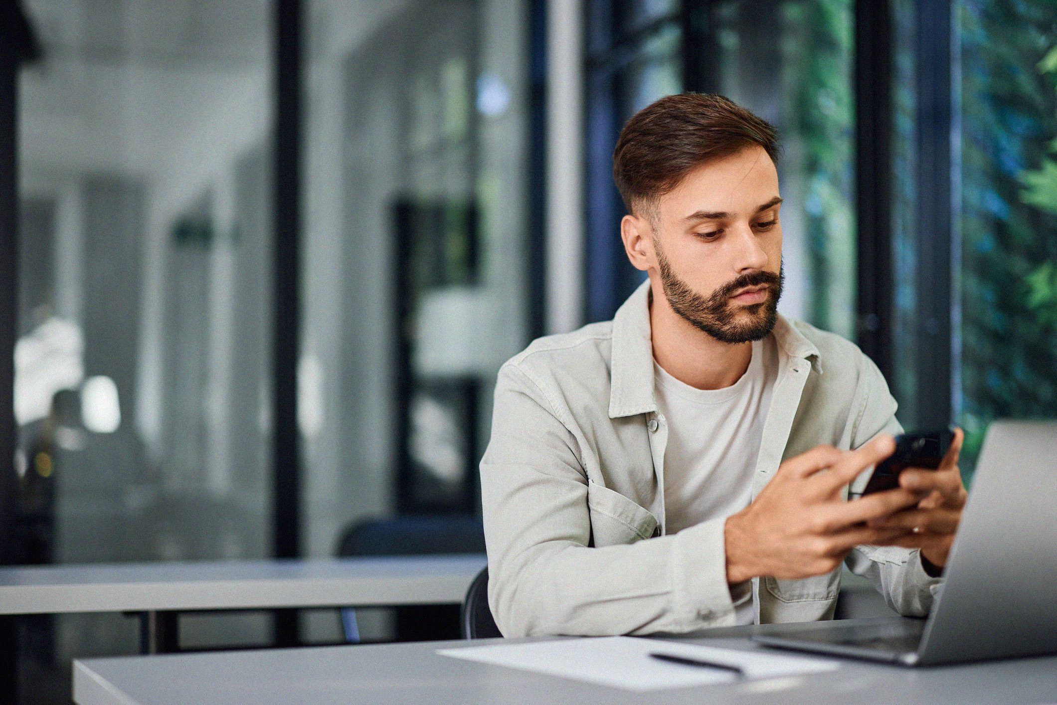 A man sitting at a desk in a modern office, looking at his smartphone while working on a laptop, with a notepad and pen in front of him.