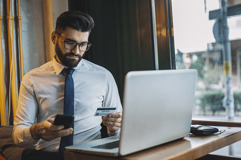 Business professional making an online payment using a credit card and smartphone at a laptop in a modern cafe setting.