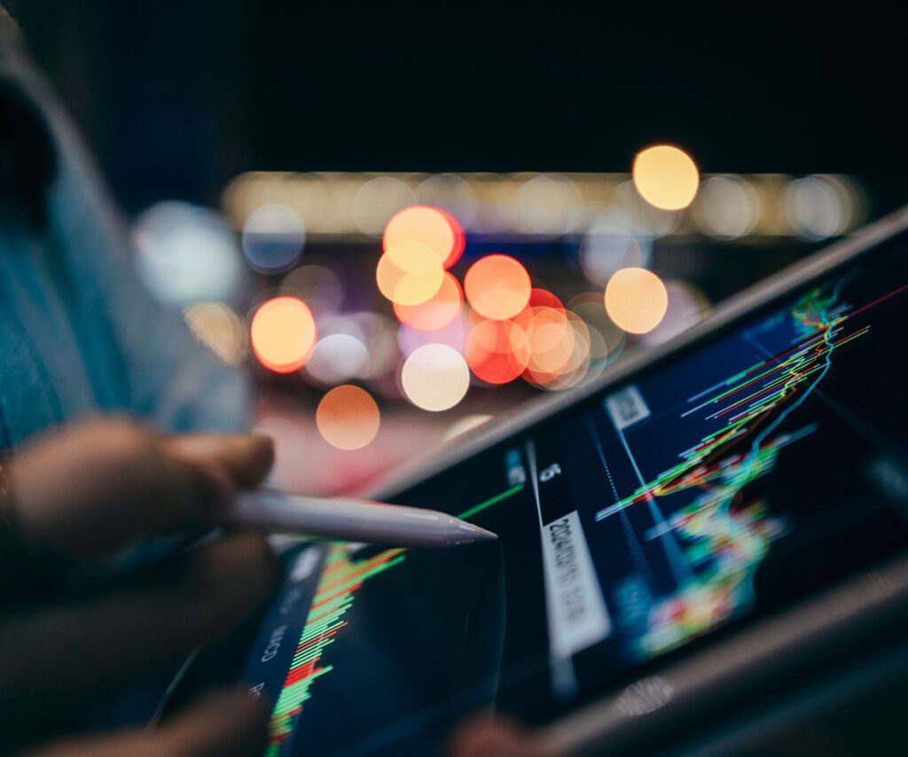Person analyzing a digital financial chart on a tablet with a stylus, against a blurred background of colorful city lights.