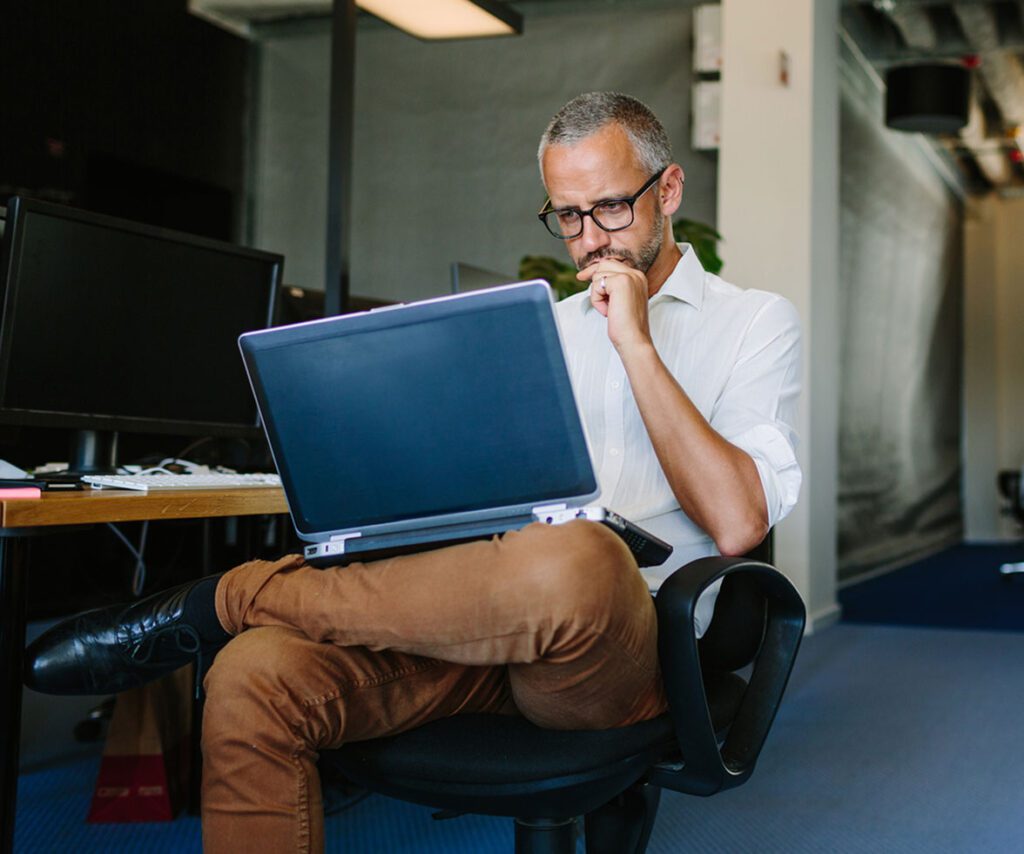 A man sitting in an office chair with his legs crossed, looking thoughtfully at a laptop in a modern office setting.