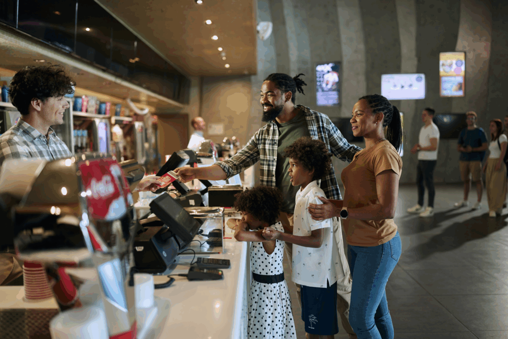 A smiling family pays at a concession stand using a card, assisted by a cashier, inside a modern sports or entertainment venue. Digital payment terminals, a Coca-Cola machine, and menu screens are visible, highlighting a streamlined payment processing experience.
