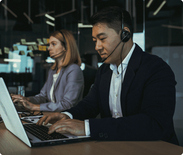 Man wearing headset working on laptop next to female coworker at a desk in an office setting