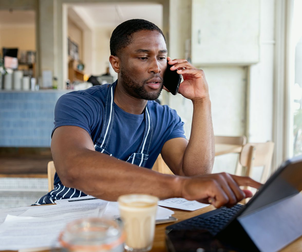 Man wearing an apron multitasking in a café, talking on the phone while working on a laptop with papers and coffee on the table.