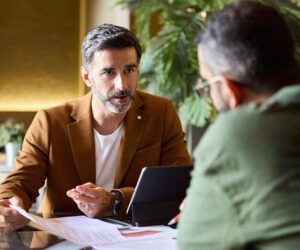 Business professional in a brown blazer discussing documents with a colleague at a desk.