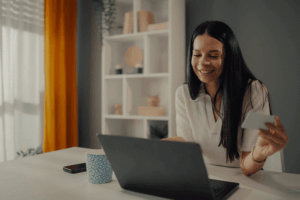 A smiling woman sits at a desk using a laptop while holding a credit card in her hand, suggesting she is shopping or making an online payment. A coffee mug and smartphone are placed on the table beside her.