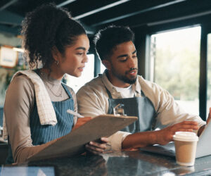 Two small business owners in aprons reviewing notes and data on a laptop together inside a café, planning operations with coffee on the counter.
