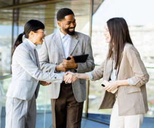 Three business professionals smiling and shaking hands during a meeting outdoors.