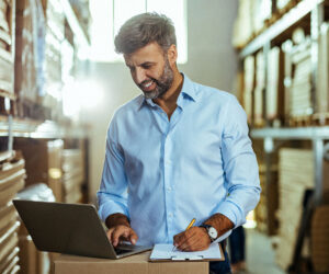 A smiling man in a light blue shirt works on a laptop and takes notes on a clipboard inside a warehouse, representing business operations or inventory management.