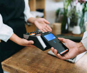 A customer makes a contactless payment by holding a smartphone near a card reader held by a cashier in a café or small shop, illustrating mobile payment technology.