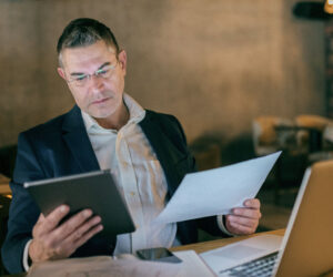 A business professional in a suit reviewing documents at a desk, holding a tablet in one hand and a sheet of paper in the other, with a laptop open in front of him in a dimly lit workspace.