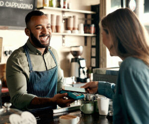 Barista smiling behind a coffee shop counter while accepting a card payment from a customer using a point-of-sale terminal.