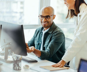 Two colleagues collaborating at a computer in a bright office, with a smiling man pointing at the monitor while a woman stands beside him.