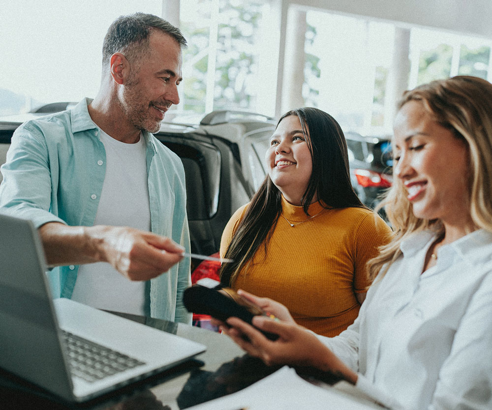 Car dealership staff reviewing a customer payment at a service counter, with a laptop and mobile device, discussing transaction details inside an auto showroom.