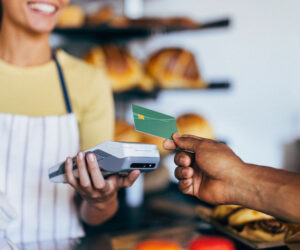 Customer handing a credit card to a cashier using a handheld card reader at a bakery counter with fresh bread in the background.