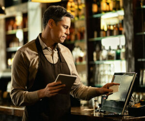 Hospitality professional operating a modern point-of-sale (POS) system in a bar setting, managing orders with a touchscreen and tablet.