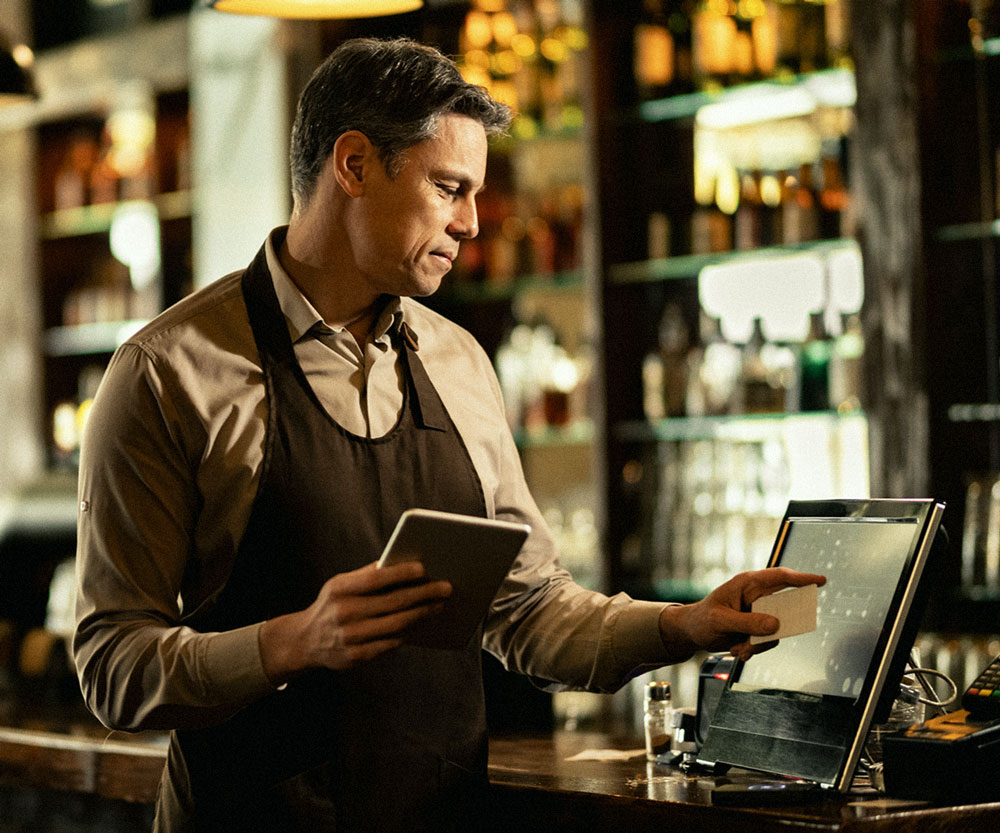 Hospitality professional operating a modern point-of-sale (POS) system in a bar setting, managing orders with a touchscreen and tablet.
