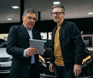 Car dealership employee showing a customer financing or payment information on a tablet while standing in a showroom with vehicles in the background.