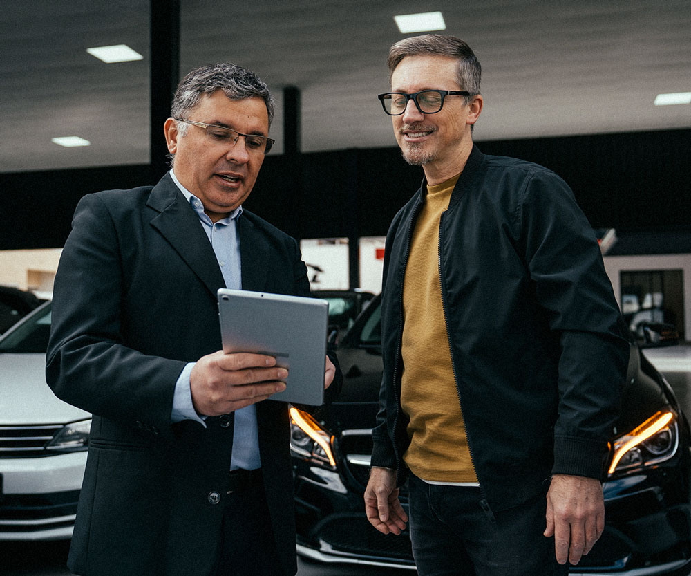 Car dealership employee showing a customer financing or payment information on a tablet while standing in a showroom with vehicles in the background.