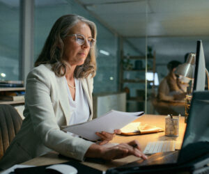 Professional woman reviewing documents while working at a desktop computer in a modern office setting.