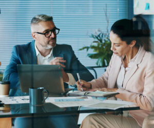 Business professionals reviewing financial documents during a meeting, with one person taking notes while another explains data on a laptop.