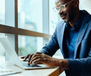 Business professional working on a laptop near a large office window, wearing glasses and a blue blazer in a modern workspace.