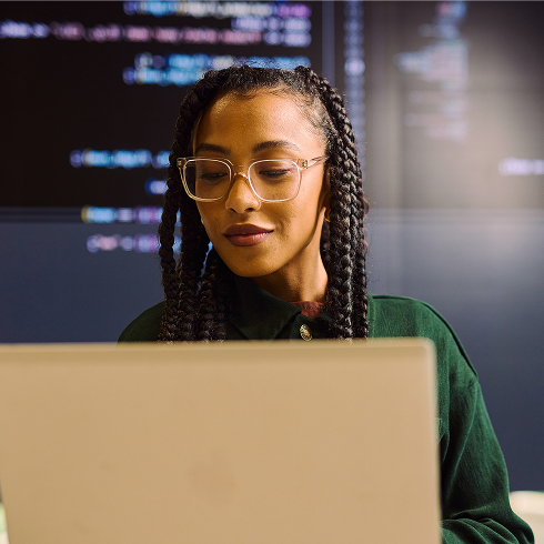 Woman with glasses and braids sitting in front of laptop with a data screen behind her