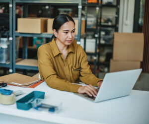 Business owner managing cash and treasury operations on a laptop in a stockroom.